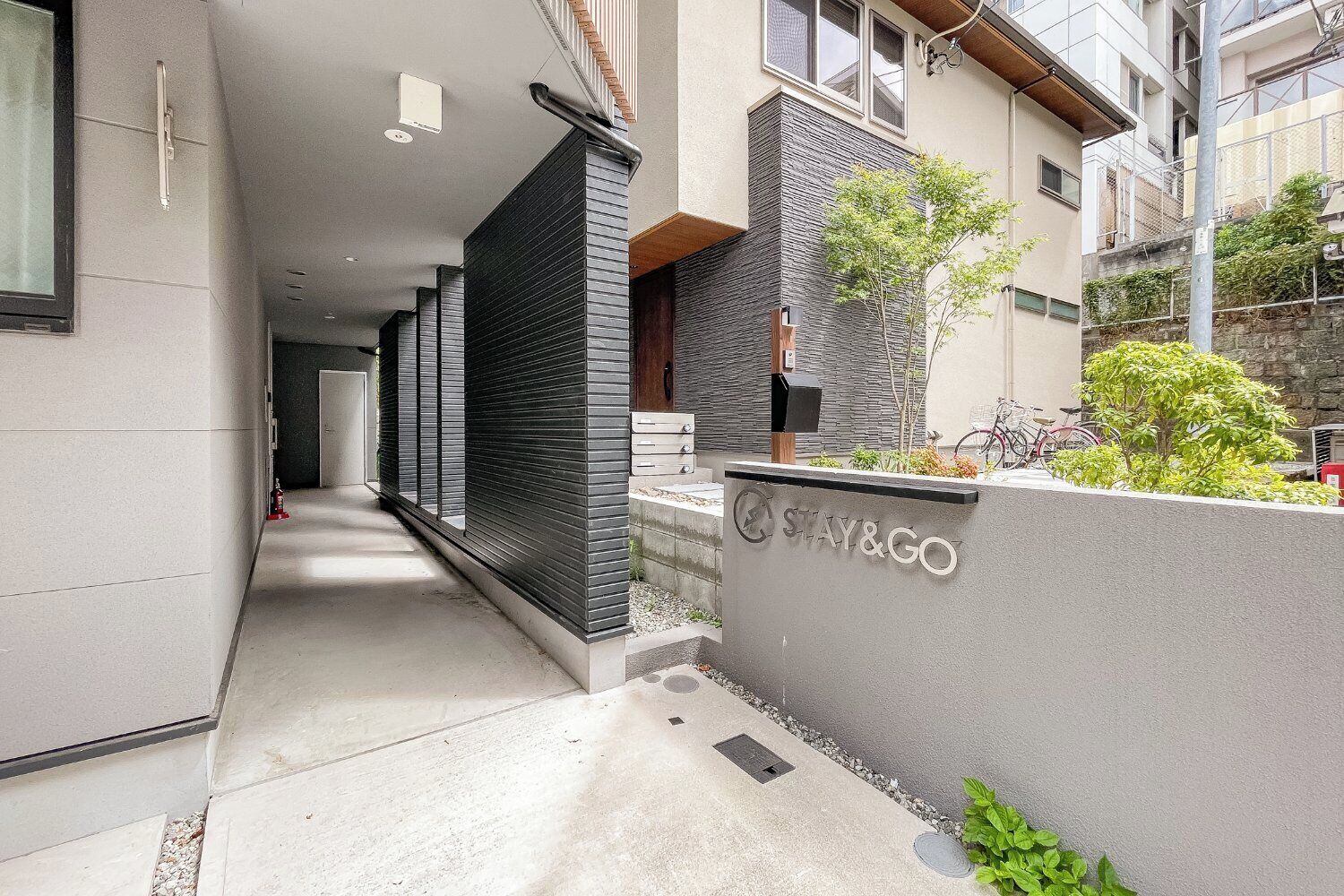 A modern building with a 'STAY&GO' sign, a narrow pathway, and a small garden area with plants and a bicycle.