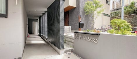 A modern building with a 'STAY&GO' sign, a narrow pathway, and a small garden area with plants and a bicycle.