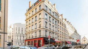 Um edifício de vários andares com entrada na esquina, toldo vermelho e vista da rua com carros estacionados.