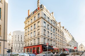 Ein mehrstöckiges Gebäude mit Hauseingang an der Ecke, einer roten Markise und einer Straßenszene mit parkenden Autos.