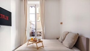 Quarto com cama, bandeja de pequenos-almoços, televisão e janela com cortinas.