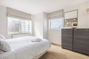 A bedroom with a bed, a dresser, a window with curtains, and a wall-mounted mirror. - Chelsea Harbour Elegance, by Veeve (London)