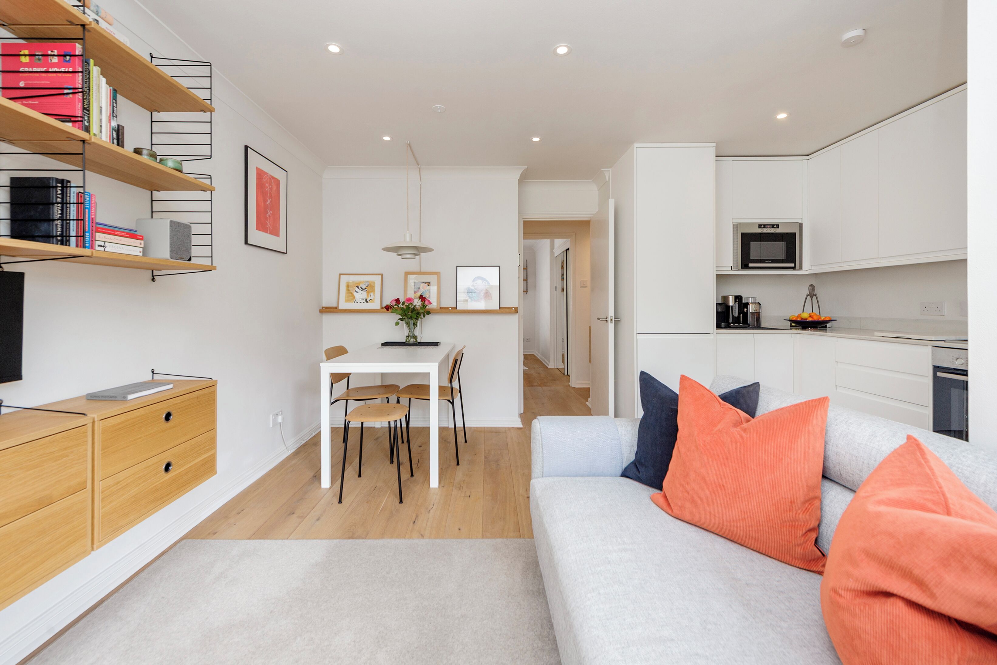 A modern apartment with a dining area, a kitchenette, a living room with a sofa and cushions, and a bookshelf with books and decorative items.