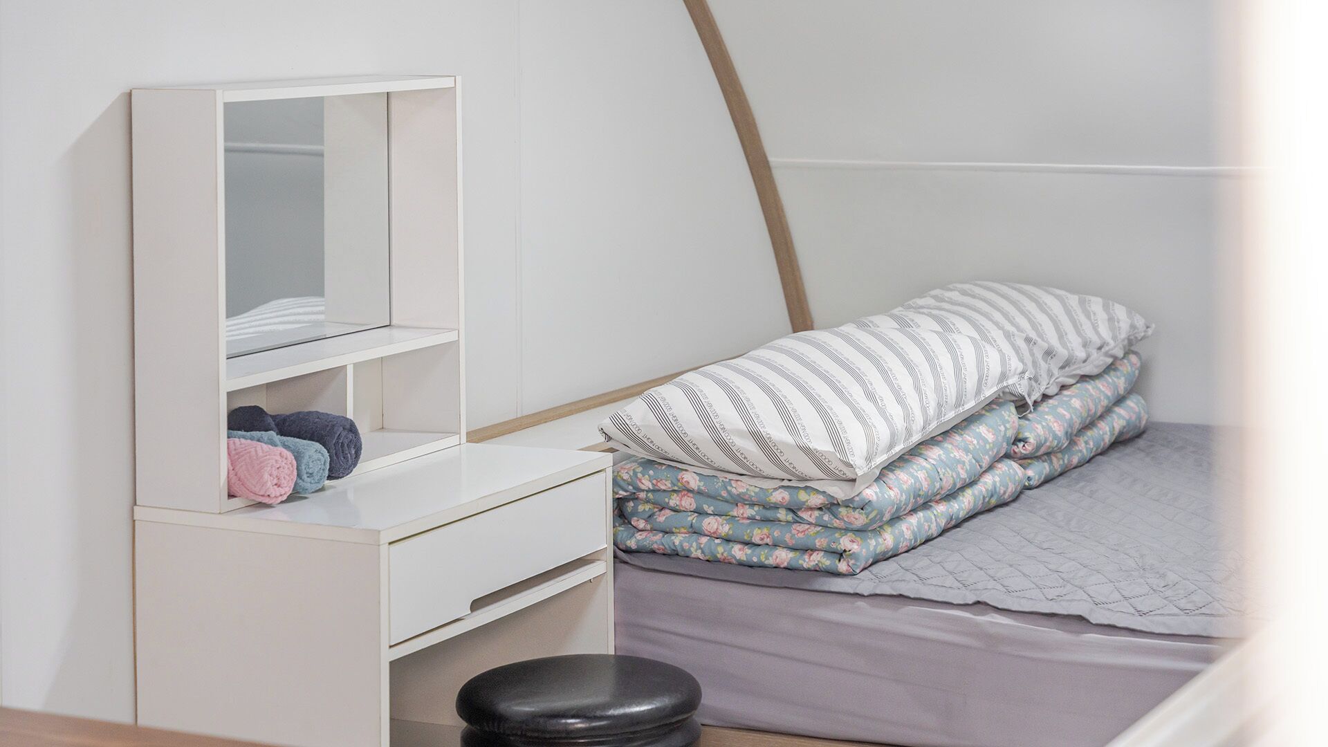 A bedroom with a white dresser, a mirror, a bed with floral bedding, and a black stool.