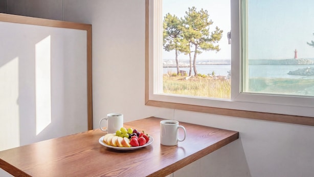 Een houten tafel met een schaal fruit en twee witte mokken, een raam met uitzicht op een vuurtoren en bomen.