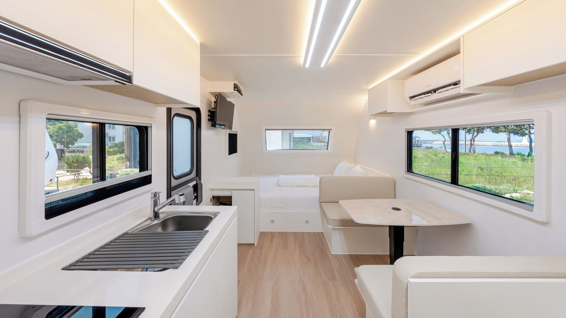 A compact RV interior with a kitchenette, a bed, and two windows offering views of the outdoors.