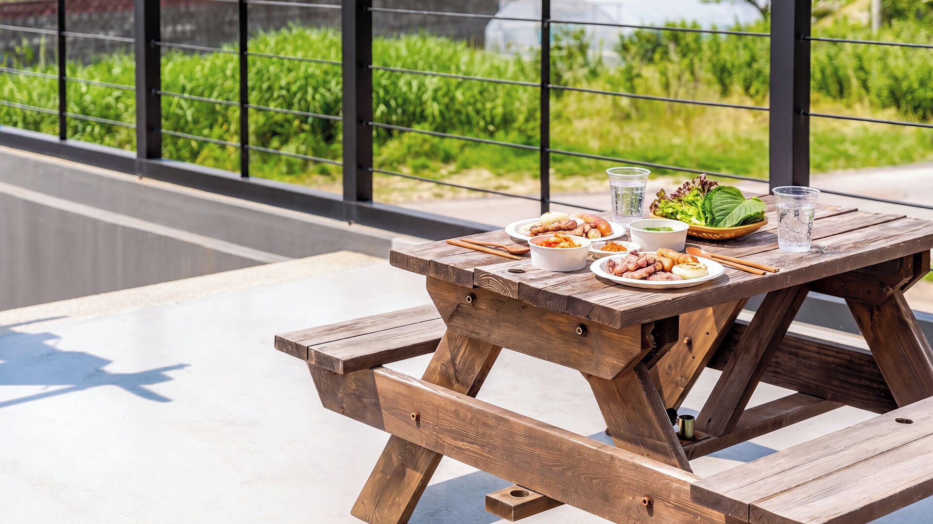 A wooden picnic table with food and drinks on it, set on a balcony with a view of greenery.