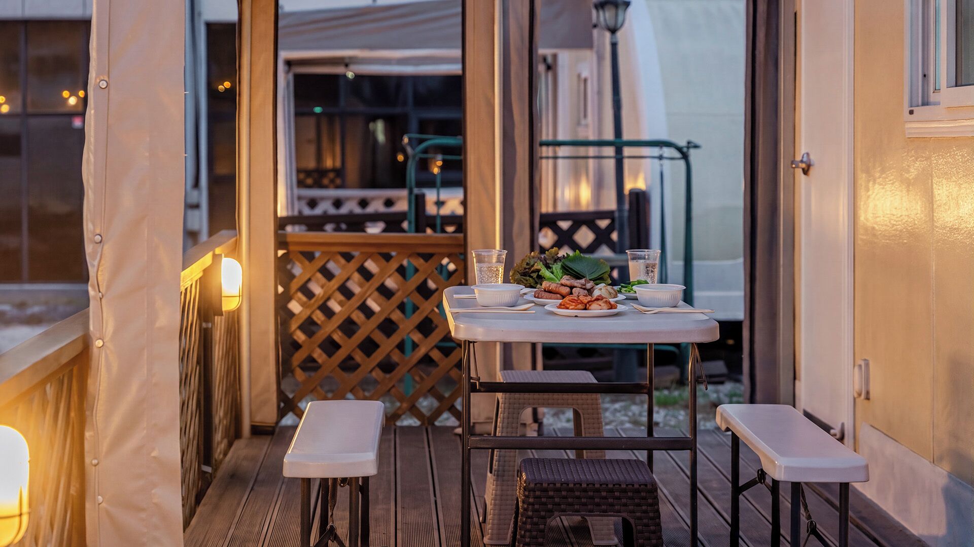 An outdoor dining area with a table set for two, a wooden deck, and a view of the building's interior.