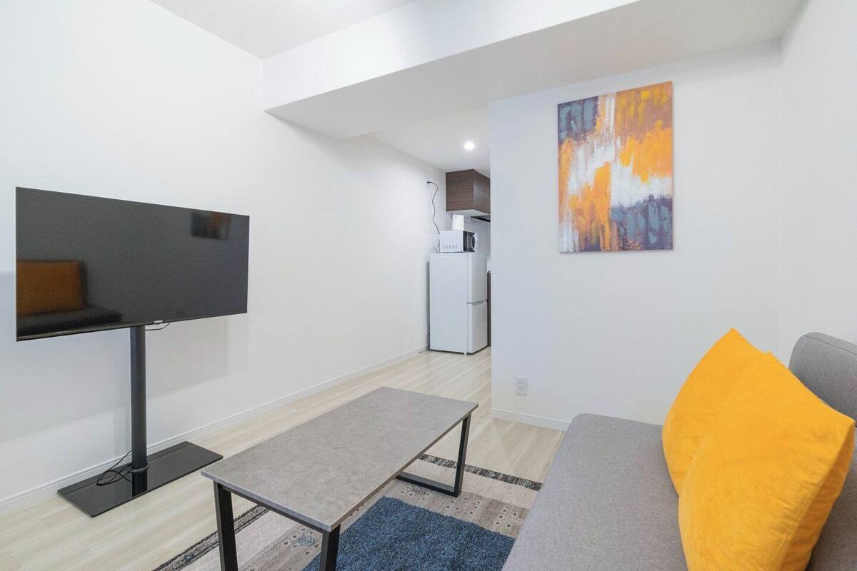 A modern living room with a flat-screen TV, a grey sofa with a yellow pillow, a small coffee table, and a kitchen in the background.