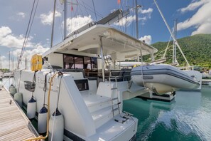 Eine Yacht liegt in einer Marina vor Anker, mit einer überdachten Deckfläche, mehreren Rettungsbooten und einer Bergkulisse im Hintergrund.