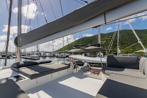 Ein Yachthafen mit vertäuten Booten, darunter ein Segelboot mit überdachter Deckfläche und Sitzgelegenheiten, sowie eine gebirgige Landschaft im Hintergrund.