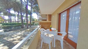 A balcony with a table and chairs, a sliding glass door, and a view of trees and a building. - Quiet Flat Surrounded by Greenery Near the Beach (Lignano Sabbiadoro)