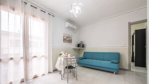 Een kamer met een blauwe bank, een tafel met een tafelkleed met bloemen, een stoel, een plafondlamp en een luchtverkoeler aan de muur.