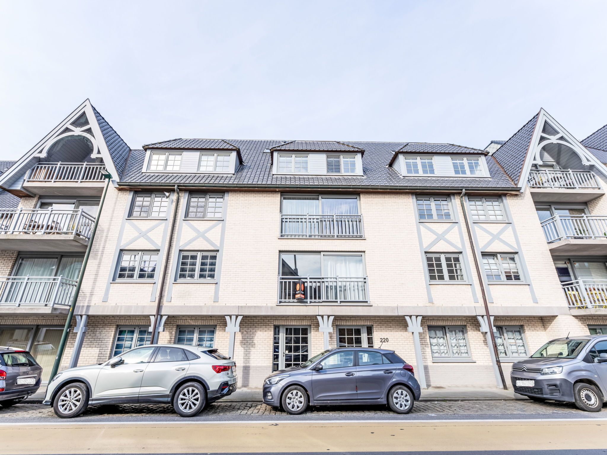 A multi-story building with a symmetrical facade, balconies, and a parking area with cars.
