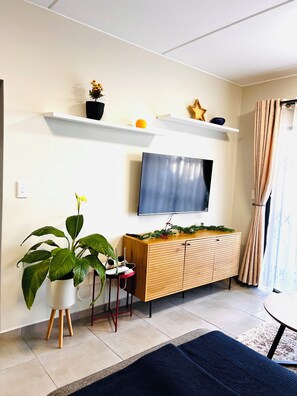 Ein Wohnzimmer mit an der Wand montiertem Fernseher, einem Holzschrank, einer Topfpflanze und einem kleinen Tisch.