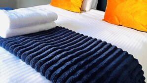 Een net opgemaakt bed met witte lakens, twee oranje kussens en een donkerblauwe deken.