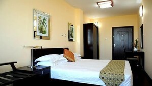 A hotel room with a bed, a nightstand, a desk, a chair, a door, and wall art. - Destiny Addis Hotel (Addis Ababa)
