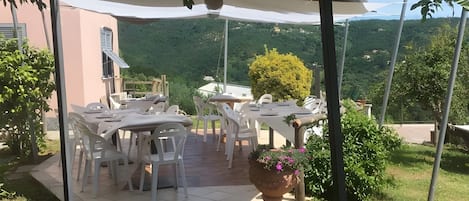 An outdoor patio with tables and chairs, a large umbrella, and a view of a mountainous landscape.
