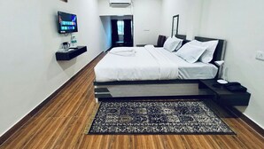 Ein Hotelzimmer mit Bett, Fernseher, Schreibtisch, Stuhl, Nachttisch, Lampe, Deckenventilator und einem gemusterten Teppich.