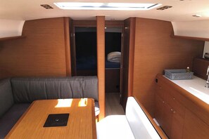 A compact interior with a wooden table, a cushioned bench, a small kitchenette, and a door leading to a sleeping area. - Sun-Kissed Sailing Adventure in the Saronic Islands (Elliniko)