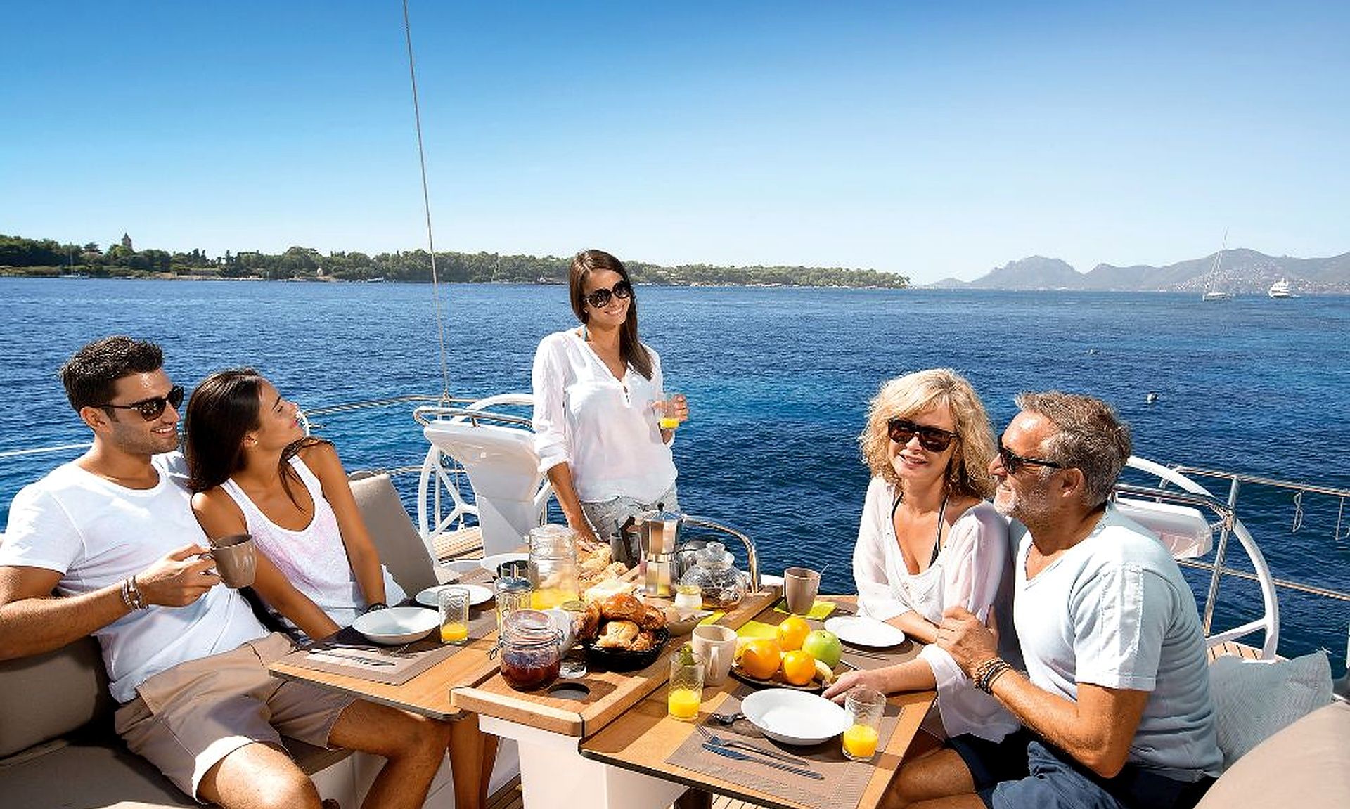 A group of people dining on a boat with a table set for breakfast, overlooking a body of water with distant land and ships.