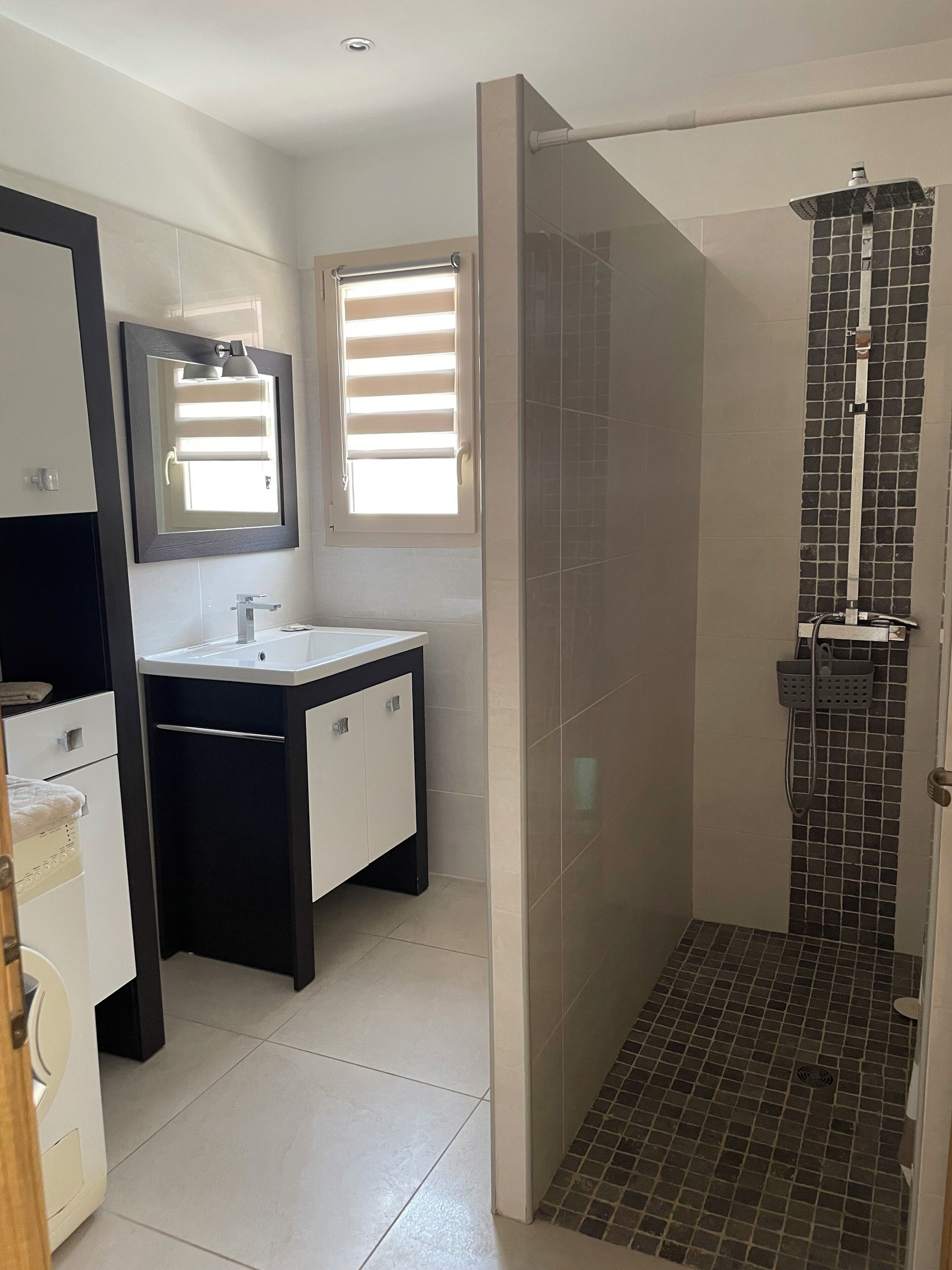 A modern bathroom with a glass-enclosed shower, a sink with a mirror, and a window with blinds.