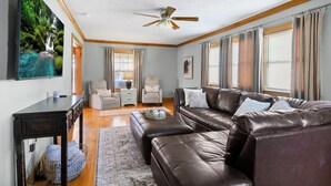 A living room with a brown leather sofa, a ceiling fan, a TV mounted on the wall, and a large window with curtains.
