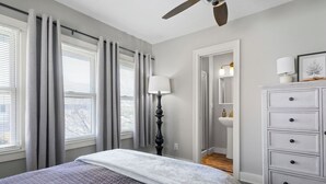 A bedroom with a bed, a dresser, a lamp, a ceiling fan, and a bathroom visible through an open door.