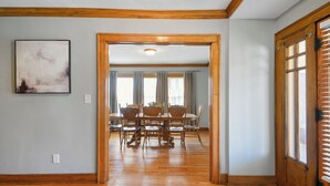 A dining area with a wooden table and chairs, a painting on the wall, and a door leading to another room.