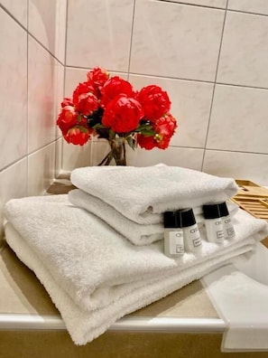 Un baño con azulejos blancos, una pila de toallas blancas y un florero con flores rojas.
