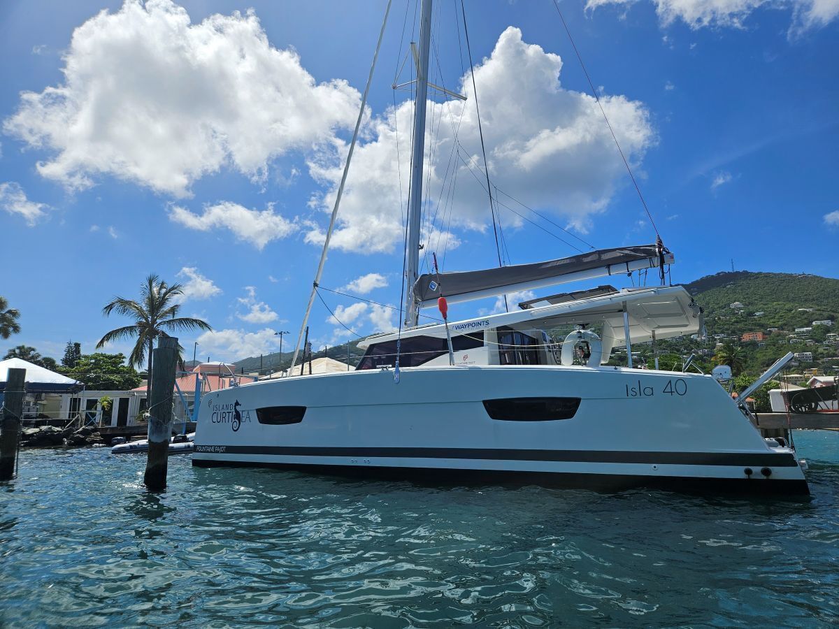 Un yate blanco llamado 'Isla 40' atracado en un puerto deportivo, bajo un cielo azul despejado y con un paisaje montañoso al fondo.