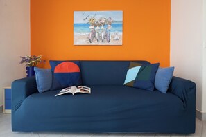 Ein blaues Sofa mit einem Buch, farbenfrohen Kissen, einer Blumenvase und einem Strandbild an einer orangefarbenen Wand.