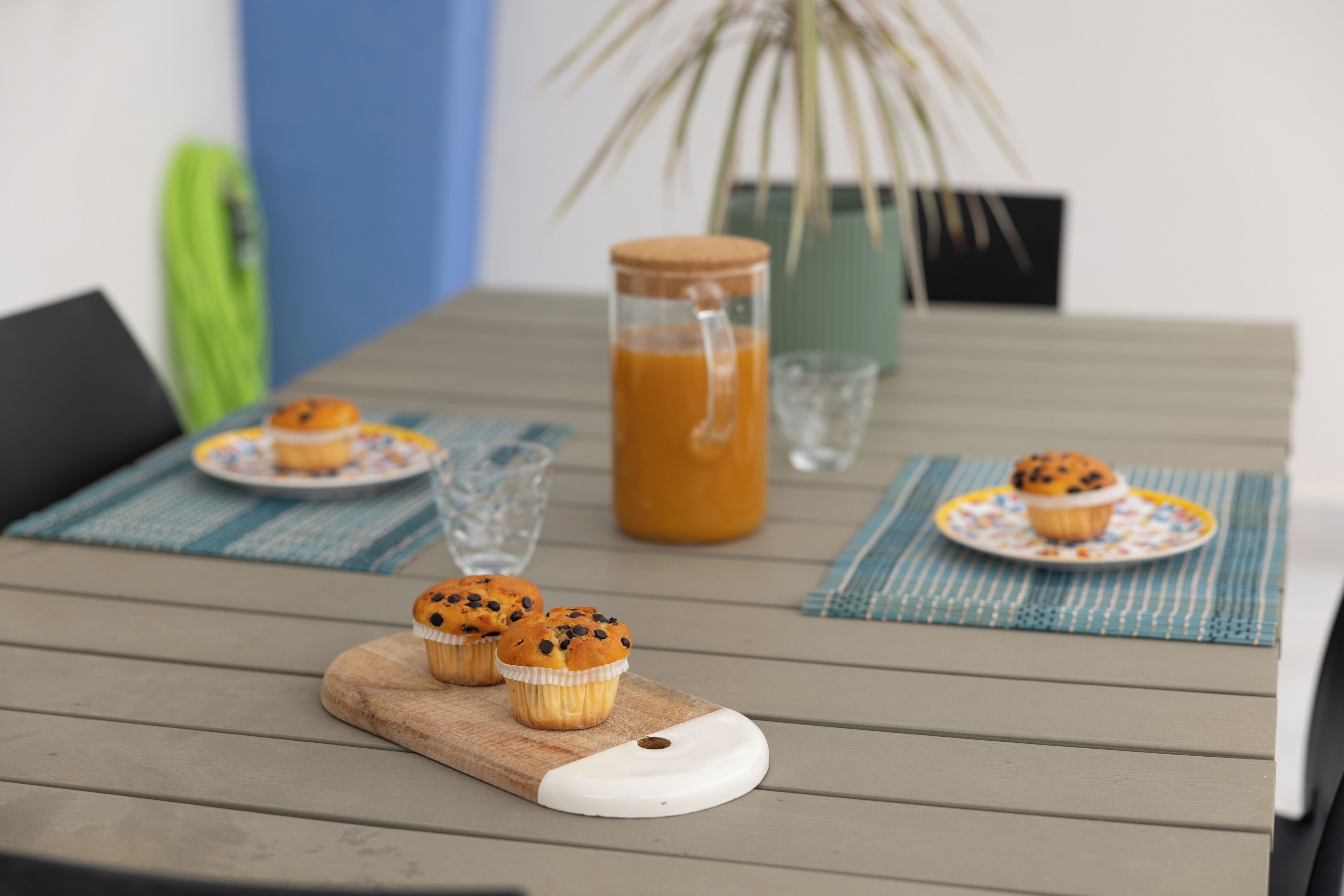 A table with a wooden cutting board, three muffins, a glass of juice, and a pitcher.
