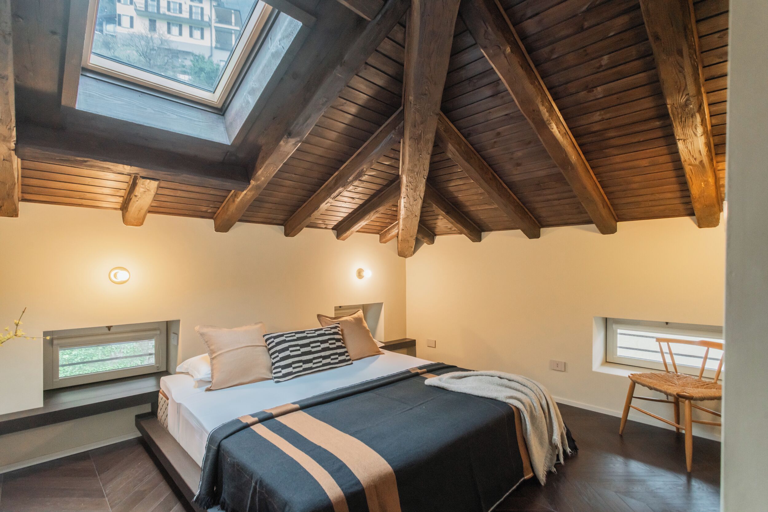 Une chambre avec un puits de lumière, un lit avec une literie à rayures, une chaise et une petite fenêtre.