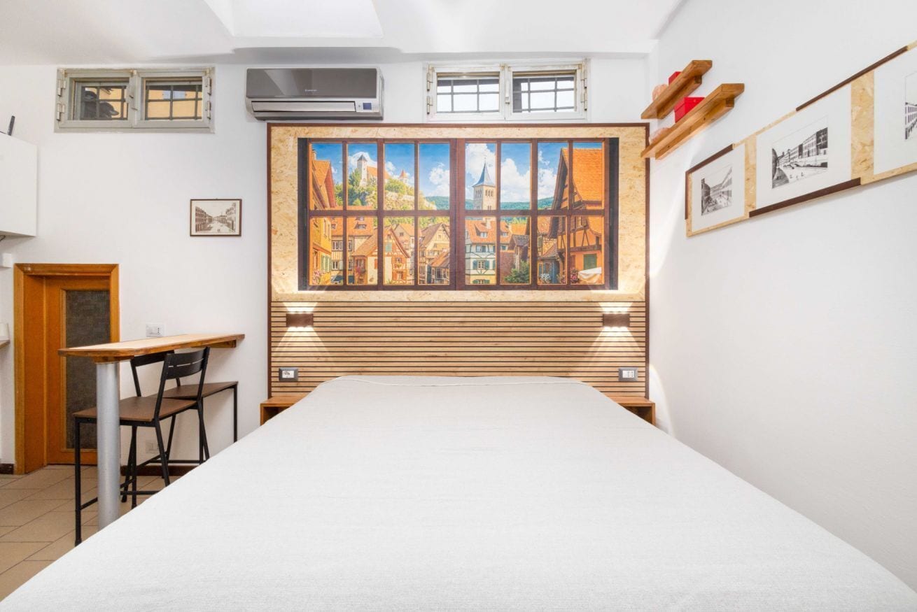 Een kamer met een bed, een bureau, een stoel, een raam met een schilderij en een luchtverwarmer aan de muur.