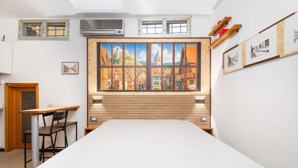 Ein Zimmer mit einem Bett, einem Schreibtisch, einem Stuhl, einem Fenster mit einem Bild und einer Wand-Klimaanlage.