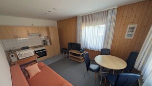 Ett kompakt rum med ett litet kök, en soffa, ett matbord med stolar och ett stort fönster med gardiner.