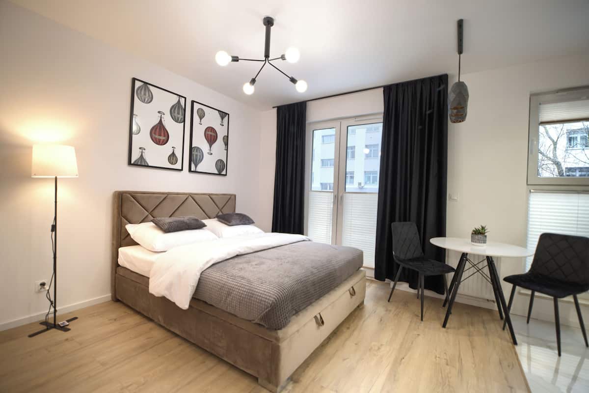Ein Schlafzimmer mit Bett, Nachttischlampe, Kunstwerk, einem Fenster mit Vorhängen und einem kleinen Tisch mit Stuhl.