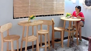 Ein Zimmer mit einem runden Holztisch, vier Holzstühlen, einem Standventilator, einer Wandklimaanlage und einem großen Fenster mit Jalousien.
