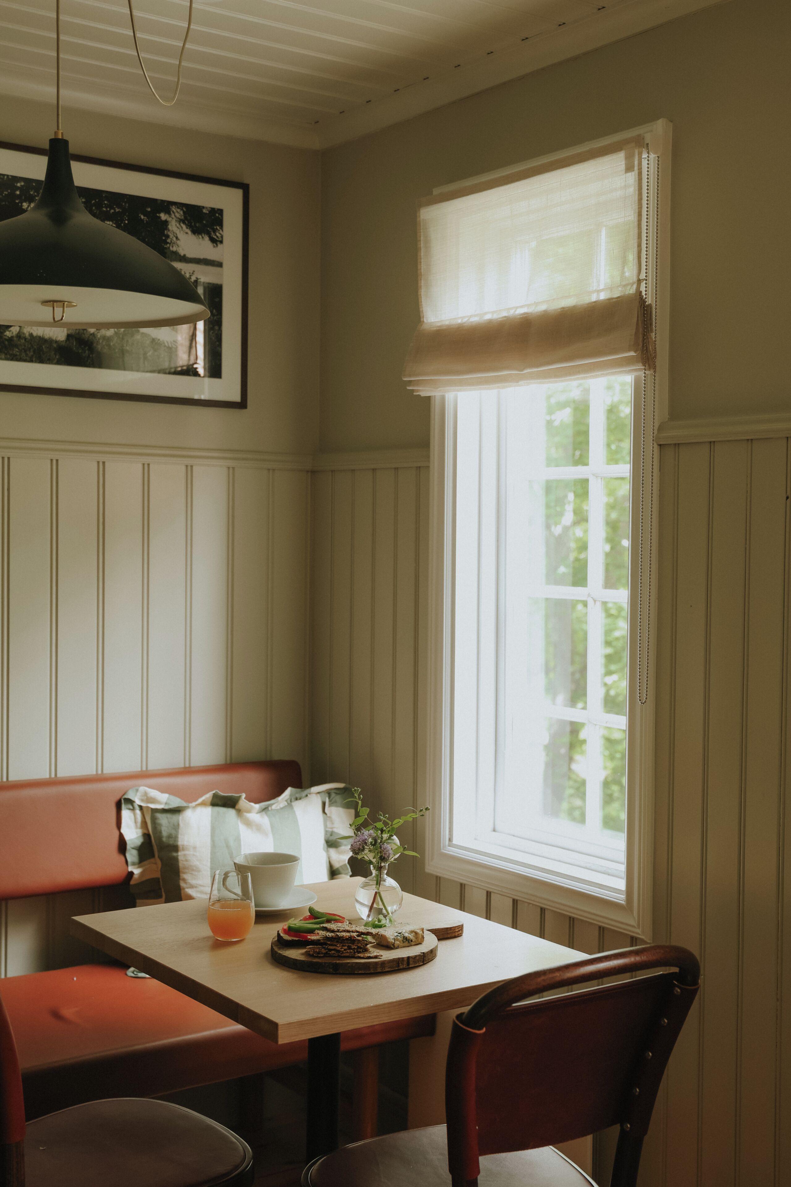 Et spiseområde med et bord, bænk og stole, et vindue med persienner samt en hængende lampe.