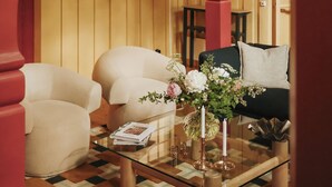 Una habitación con una mesa de centro de vidrio, dos sillones color crema, un sofá verde oscuro y una columna roja.