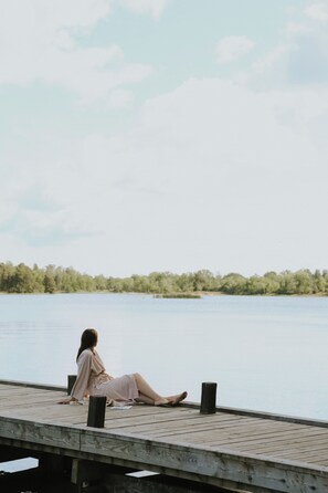 A woman sitting on a wooden dock by a calm lake, with trees in the background. - Badhotellet Villa Berg  (Norrtalje)