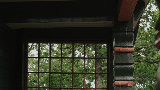 Una persona sentada en una silla de mimbre junto a una ventana con vistas a árboles.