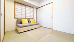 Ein Zimmer mit Tatami-Boden, einer niedrigen Holzliege mit Kissen, einer verschiebbaren Shoji-Schirmwand und einer traditionellen japanischen Schiebetür.