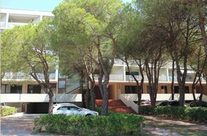 A building with a modern architectural style, featuring a staircase leading to an upper level, surrounded by trees and parked cars. - Practical Flat Just a Short Walk From the Beach (Lignano Sabbiadoro)