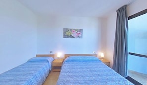 A hotel room with two beds, a nightstand with a lamp, a painting on the wall, and a window with curtains. - Practical Flat Just a Short Walk From the Beach (Lignano Sabbiadoro)