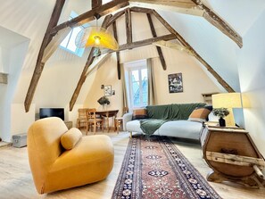 Ein Loft-Zimmer mit freiliegenden Holzbalken, einer Couch, einem Sessel, einem Tisch mit Stühlen und einem gemusterten Teppich.