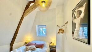 Un dormitorio con techo inclinado, una cama con almohadas, una lámpara colgante, un ventanal pequeño y un cuadro en la pared.