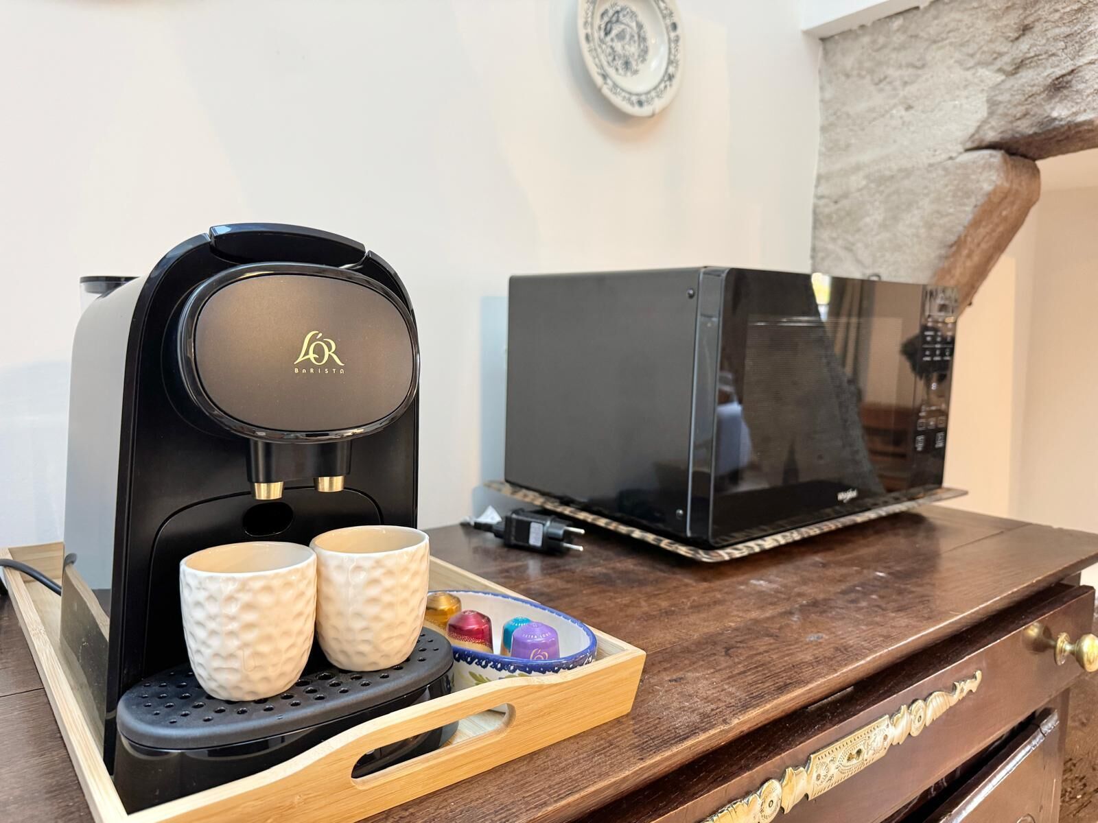 Una cafetera con dos tazas, una bandeja con bombones y un microondas sobre una mesa de madera.