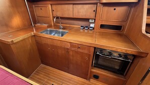 A compact galley kitchen with a stainless steel sink, wooden cabinets, a built-in oven, and a window above the sink. - Sun-Kissed Sailing Adventure in Cataluña (Volos)